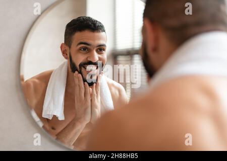 Ragazzo arabo fiducioso giovane che guarda nello specchio, toccando la barba Foto Stock