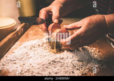 Donna anziana aggiungere l'uovo alla farina. Preparare l'impasto su tavola di legno. Vecchia donna che cucina in cucina domestica. Immagine a toni Foto Stock