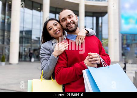Coppia interrazziale affettuosa con borse shopper sorridenti e abbracciate vicino al grande centro commerciale, all'aperto Foto Stock