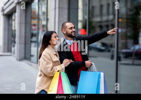 Coppia interracial positiva camminando lungo la strada con i sacchetti di carta, indicando alla nuova finestra del negozio, shopping insieme Foto Stock