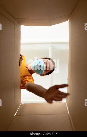 Giovane donna asiatica in maschera medica sboxing pacco di cartone, cercando di ottenere qualcosa, vista dall'interno Foto Stock