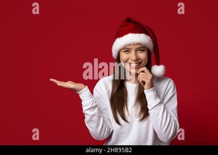 Offerta natalizia. Giovane donna allegra in Santa Hat che tiene invisibile oggetto su palma aperto Foto Stock