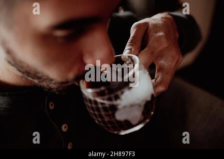 Il giovane uomo misto sorsegge il caffè nero dalla tazza di novità alla luce del mattino. Primo piano bere caffè caldo da una tazza di vetro. Pausa caffè durante la giornata lavorativa Foto Stock