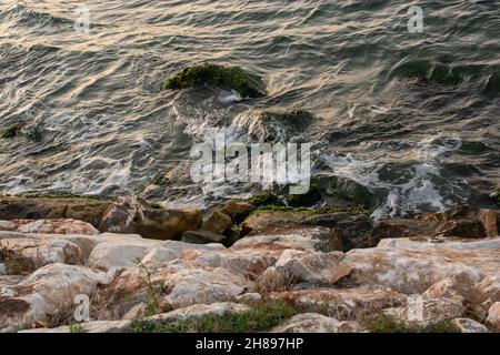 Una bella onde schiumose che si infrangono su rocce di costa ruvida e musciose Foto Stock