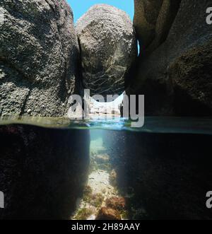 Stretto passaggio sotto un masso incastonato tra rocce sulla riva del mare, vista divisa sopra e sotto la superficie dell'acqua, Spagna, Galizia, provincia di Pontevedra Foto Stock