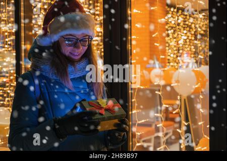 Graziosa Donna in cappello Santas con scatola di regali vicino alla finestra del caffè illuminato. Lei ondeggia la mano e attende il suo amato compagno. Natale presenta vacanze, Foto Stock