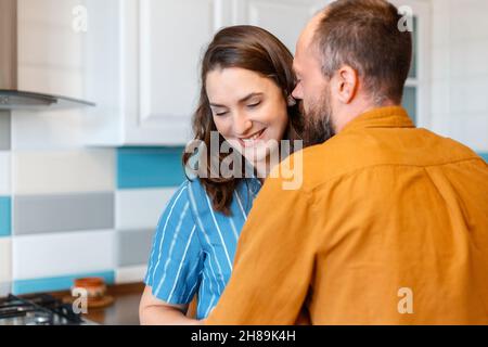 Felice giovane amorevole coppia uomo donna 30s in cucina a casa. Sposata moglie di coppia e marito che abbracciano divertirsi sorridendo bacio in cucina interno di Foto Stock