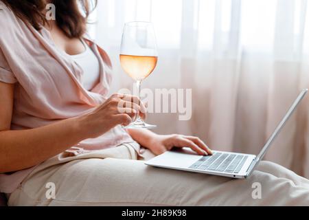 Giovane bella donna che beve vino di rosa e l'uso del computer portatile. Donna caucasica rilassata guardando il film su laptop o effettuare acquisti online. Laptop e vetro Foto Stock