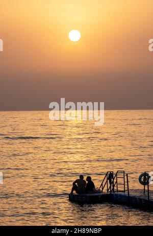 Una coppia guarda il tramonto mentre si siedono insieme su un pontile a Paphos, Cipro. Foto Stock