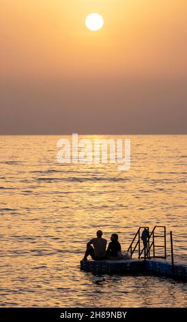 Una coppia guarda il tramonto mentre si siedono insieme su un pontile a Paphos, Cipro. Foto Stock