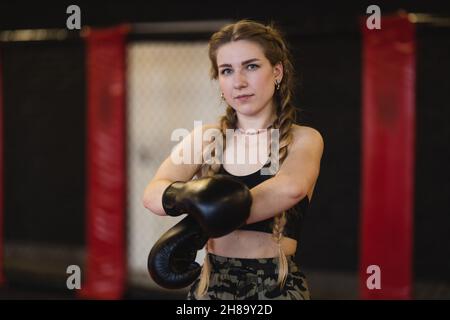 Allenamento fitness boxe femminile in palestra. Giovane donna con trecce si prepara per un allenamento, indossa guanti da boxe e guarda una macchina fotografica. Foto Stock