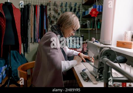 Una donna dressmaker utilizza una macchina da cucire per creare abiti personalizzati in un'officina. Una sartstress femminile è occupato cucire abiti sul posto di lavoro. Foto Stock