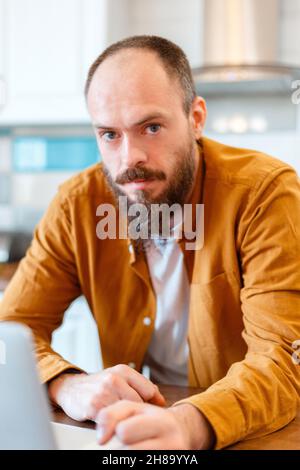 Ritratto calma uomo che lavora al computer portatile in cucina a casa. Bald bearded 30 uomo guardando la telecamera Foto Stock