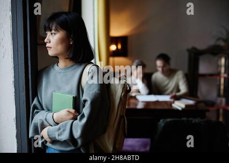 Giovane donna pensiva con libri che guardano attraverso la finestra mentre si alza nella stanza Foto Stock