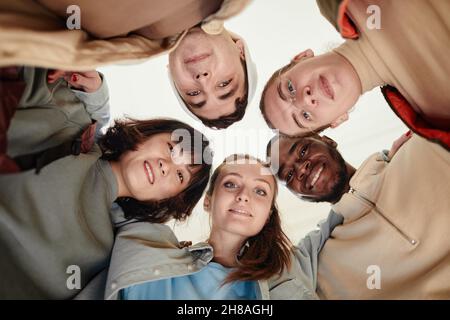 Vista ad angolo basso degli adolescenti che si abbracciano mentre sono in piedi nel cerchio e guardano la fotocamera Foto Stock