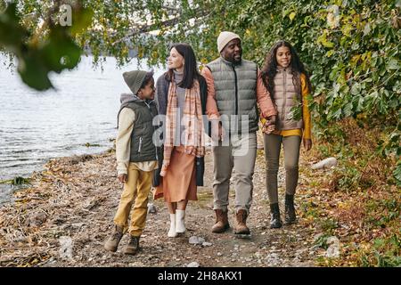 Felice giovane famiglia interrazziale di genitori e due bambini che si muovono lungo il litorale mentre godendo a piedi il giorno d'autunno Foto Stock