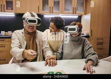 Carino ragazzo africano che guarda il nonno in visore vr premendo il tasto del telecomando mentre si trova tra i nonni Foto Stock