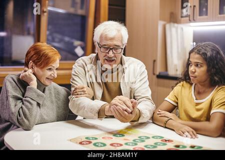 L'uomo maturo che scuote i dadi nelle mani sopra il gioco del bordo mentre lo gioca con la sua moglie e la loro nonna interracial cute Foto Stock