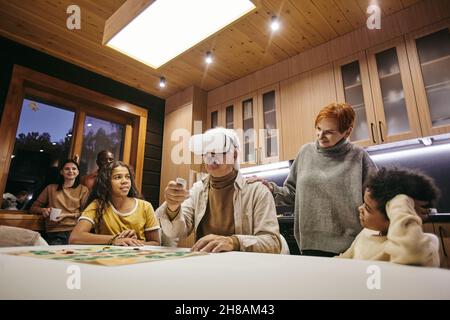 Donna matura e i suoi due nipoti interrazziali che guardano l'uomo anziano in cuffie per realtà virtuale durante il gioco di piacere Foto Stock