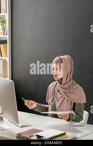 Giovane insegnante femminile in hijab seduta di fronte al computer e spiegare l'argomento della lezione al pubblico online Foto Stock