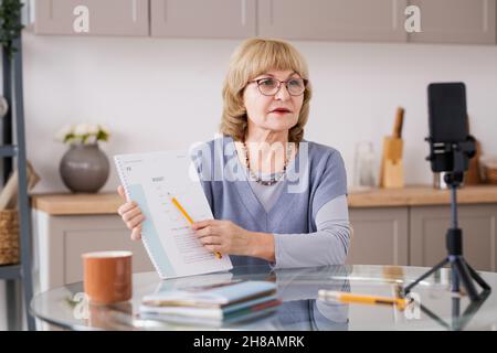 Insegnante anziano bionda femminile che punta al documento finanziario mentre si siede dal tavolo da cucina di fronte alla fotocamera dello smartphone Foto Stock