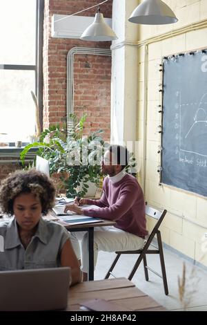Giovane uomo d'affari seduto sul posto di lavoro di fronte al monitor del computer, mentre la sua collega femminile digita e guarda il display del notebook Foto Stock