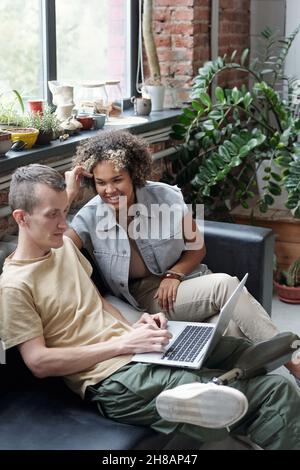 Felice giovane interculturale donna che guarda il collega maschio con il laptop durante la discussione dei punti di lavoro in riunione Foto Stock