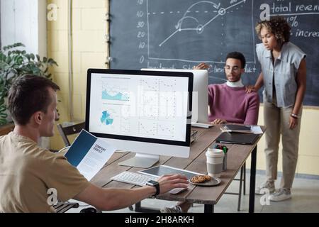 Giovane economista che guarda i dati finanziari sullo schermo del computer mentre si siede di fronte ai colleghi che discutono di informazioni online Foto Stock