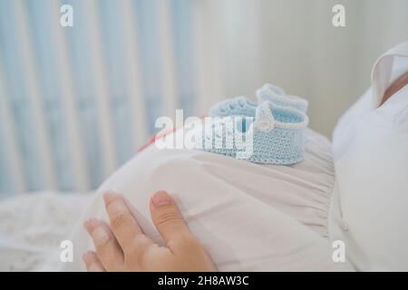 Crop donna incinta senza volto con scarpe per bambini sul pancino che giace sul letto Foto Stock