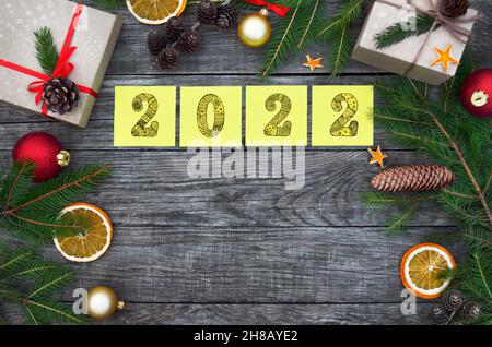 Note gialle adesive con numeri 2022 su sfondo festoso in legno con spazio per la copia. Composizione anno nuovo con scatole regalo, rami di abete, coni e de Foto Stock