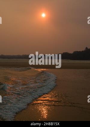 Tramonto arancio fumoso sulle onde che si infrangono sulla spiaggia, riflesso del sole sulla sabbia bagnata Foto Stock