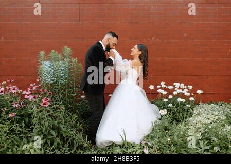 Lo sposo bacia la mano contro muro di mattoni e fiori Foto Stock