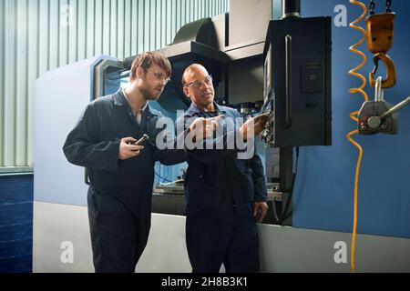 Un lavoratore esperto spiega il funzionamento di una macchina CNC per un apprendista Foto Stock