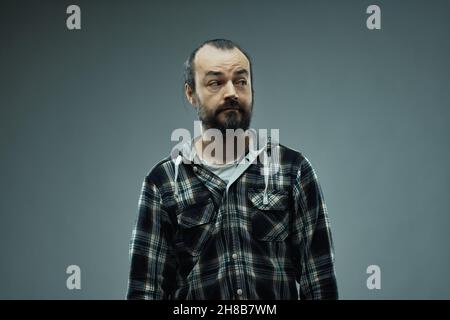 Ritratto da studio di un uomo con bearded che indossa una camicia a scacchi guardando a sinistra con un'espressione facciale sospetta su sfondo grigio per c Foto Stock