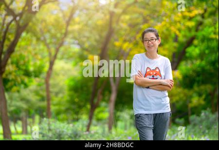 Ritratto di donna asiatica di mezza età in posa in un parco con abiti casual per l'esercizio fisico, in piedi con le braccia a croce sul petto, sfondo sfocato di alberi Foto Stock