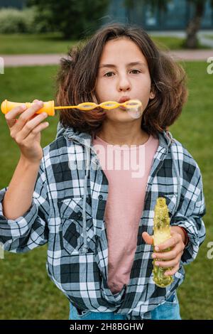 Attractiveschoolgirl di etnia caucasica fa bolle di sapone. Ritratto di una bella bruna ragazza carina divertirsi in un parco estivo. Emotini positivi Foto Stock