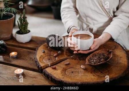 Cacao cerimoniale caldo fatto a mano in tazza bianca. Donna mani che tengono cacao artigianale, vista dall'alto su tavola di legno. Bevanda biologica sana al cioccolato preparata da Foto Stock