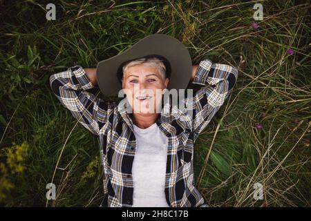Vista dall'alto della donna anziana felice agricoltore che si trova sull'erba e guarda la macchina fotografica all'aperto nel prato. Foto Stock