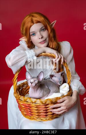 Cospayer elfo giovane donna rossa sorride, appoggiandosi su un cestino marrone in cui due simpatici cuccioli di Sphinx sono seduti con calma. Elf in abito bianco, con capelli Foto Stock