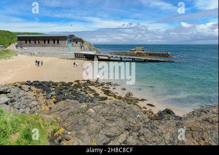 Stazione di salvagente, scivoli e molo in una tranquilla giornata estiva con cielo e mare blu; persone che camminano sulla spiaggia con sentiero erboso e rocce in primo piano. Foto Stock