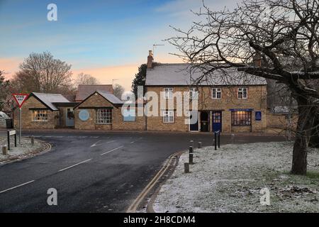 Sharnbrook High Street, Bedfordshire, Inghilterra, Regno Unito - scena invernale all'alba di negozi e cottage sul verde villaggio con gelo e neve sui tetti Foto Stock