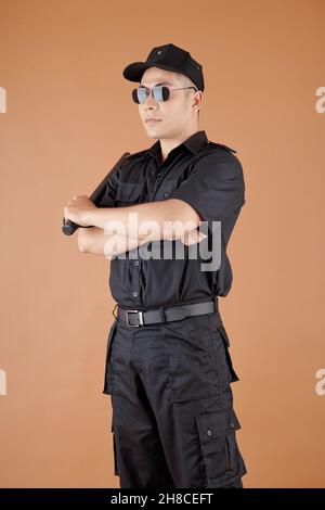 Giovane ufficiale di polizia serio in occhiali da sole e uniforme tenendo batone e guardando via Foto Stock