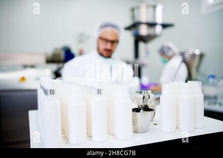 Giovane tecnologo che fa creme per la cura del viso. Seduto in una fabbrica di prodotti cosmetici che lavora con un computer portatile. Foto Stock