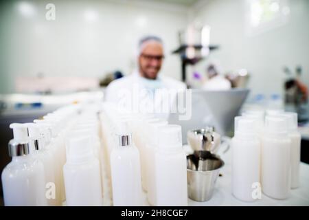 Giovane tecnologo che fa creme per la cura del viso. Seduto in una fabbrica di prodotti cosmetici che lavora con un computer portatile. Foto Stock