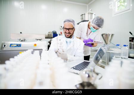 Giovane tecnologo che fa creme per la cura del viso. Seduto in una fabbrica di prodotti cosmetici con la sua assistente femminile. Foto Stock