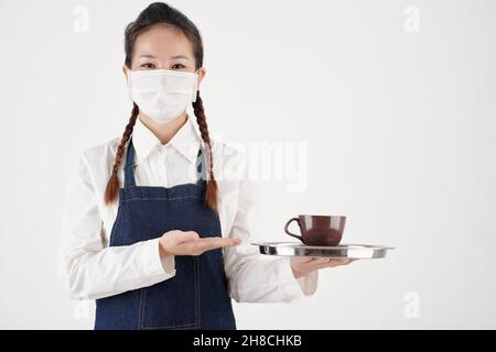 Ritratto di cameriera caffè in maschera medica che serve una tazza di buon caffè al cliente Foto Stock