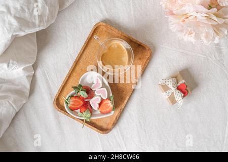 Colazione per San Valentino. Piatto bianco a forma di cuore con fragole fresche, tazza di caffè, regalo e fiori con regalo a letto. Vita morta Foto Stock