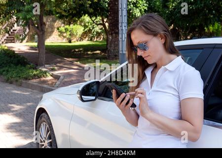 una ragazza in un abito bianco si trova vicino a una macchina bianca costosa e comunica al telefono, scrive messaggi di testo o tramite messaggeri in social network, pe Foto Stock