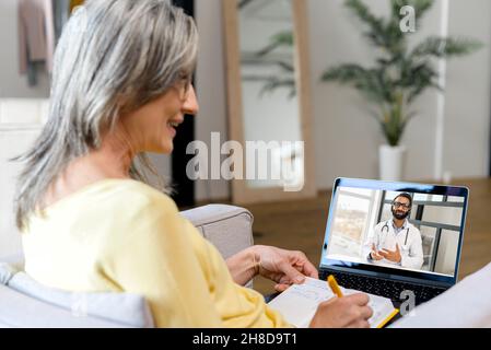 Donna matura che parla online da casa con un medico, avendo un controllo medico virtuale con lo specialista medico su una webcam, ascolta attentamente e prendendo appunti. Cyber medicina e rimanere a casa Foto Stock