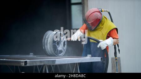 Vista laterale del lavoratore in speciale vestito protettivo e maschera che lavora o lucidando metallo per creare recinzione in smithy. Concetto di processo che rende nuova e moderna recinzione. Foto Stock